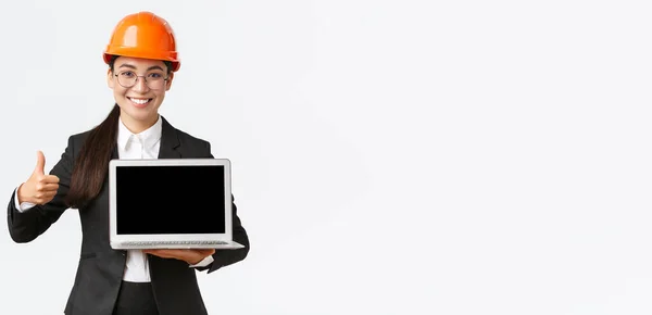 Uśmiechnięta profesjonalna architekt, kierownik budowy w fabryce pokazuje wykres, pozytywny diagram, kciuki do góry w zatwierdzeniu i trzymając laptop skierowany do ekranu, nosić kask bezpieczeństwa — Zdjęcie stockowe