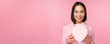 Bu da senin için. Romantik, sevimli Asyalı şirket kadını, takım elbiseli kız, kalp şeklindeki kutuyu hediyeyle gösteriyor, pembe arka planda duruyor.