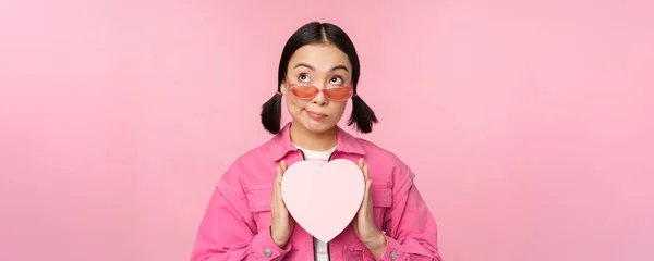 Schöne asiatische Mädchen lächelt glücklich, zeigt Herz Geschenkbox und schaut aufgeregt in die Kamera, steht über rosa romantischen Hintergrund — Stockfoto