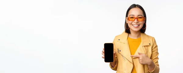 Porträtt av snygg koreansk tjej i solglasögon, leende, pekar finger åt smartphone skärm, visar mobiltelefon ansökan, står över vit bakgrund — Stockfoto