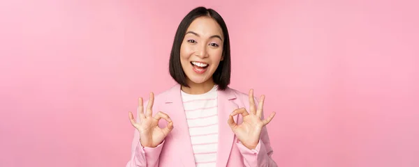 Oké, uitstekend. Zakenvrouw in corporate suit, tonen ok, goedkeuring gebaar, aanbevelen smth, geven positieve feedback en glimlachen blij, poseren over roze achtergrond — Stockfoto