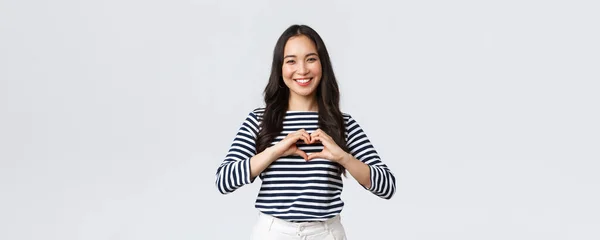 Lebensstil, Menschen Emotionen und lässiges Konzept. Schöne lächelnde entzückende asiatische Frau, die Herzzeichen zeigt und lächelt, Sympathie oder Fürsorge ausdrückt, weißer Hintergrund stehend — Stockfoto