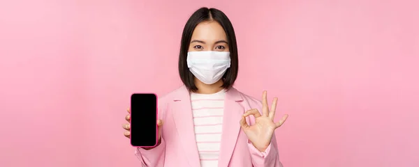 Portrait of asian businesswoman in medical face mask covid-19, showing smartphone screen and okay sign, pink background — Stock Photo, Image