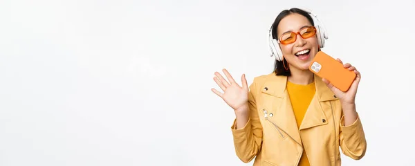 Felice donna asiatica elegante in occhiali da sole, ascoltare musica in cuffia, tenendo smartphone, canto e danza, in piedi su sfondo bianco — Foto Stock