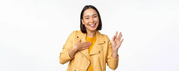 Immagine di amichevole ragazza asiatica in elegante cappotto giallo, alzando il braccio, presentarsi, saluto, agitando la mano, dicendo ciao, in piedi su sfondo bianco — Foto Stock