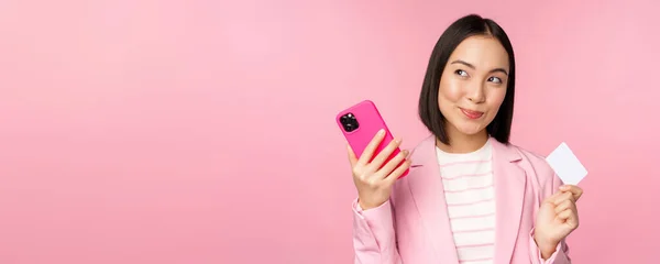 Sorrindo mulher corporativa asiática, senhora de terno pensando, segurando smartphone e cartão de crédito, plano para comprar smth on-line, compras com telefone celular, fundo rosa — Fotografia de Stock