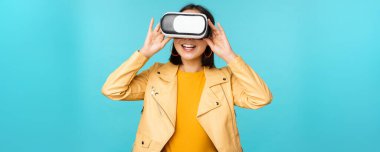 VR kulaklık takan mutlu Asyalı kadın modelin portresi. Sanal gerçeklik gözlükleriyle gülüyor ve gülümsüyor. Mavi arka planda duruyor.