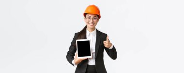 Profesyonel gülümseyen Asyalı kadın inşaat yöneticisi, iş elbisesi içindeki fabrika mühendisi ve güvenlik kaskı olumlu grafikli dijital tablet ekranı, beyaz arka plan