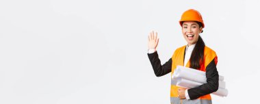 Dost canlısı, neşeli Asyalı kadın inşaat yöneticisi, güvenlik kaskı ve ceketli baş mimar, bina projesinin planları, el sallayarak merhaba demek, birini selamlamak,