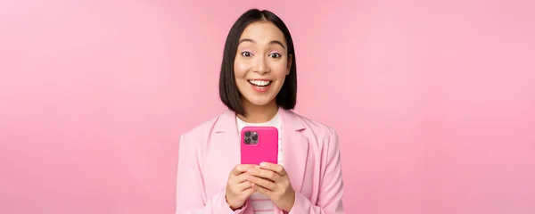 Portré ázsiai lány öltönyben okostelefon, mosolygós és boldognak tűnik, felett áll rózsaszín stúdió háttér — Stock Fotó