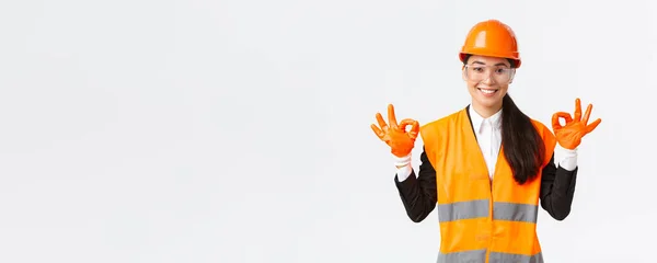 Αυτοπεποίθηση ασιατική γυναίκα μηχανικός κατασκευών, διευθυντής επιχείρησης δείχνει εντάξει χειρονομία μετά τη θέση σε κράνος ασφαλείας, γυαλιά και γάντια πριν από την είσοδο σε επικίνδυνη περιοχή, λευκό φόντο — Φωτογραφία Αρχείου