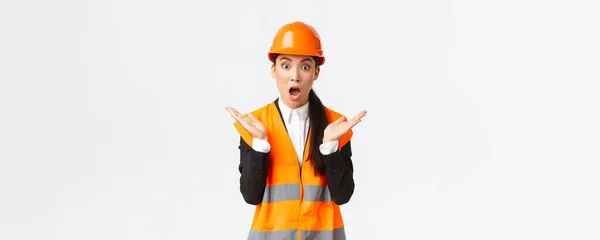 Surprised and shocked asian female engineer in reflective jacket, safety helmet, react to unexpected news, raising hands up and clap impressed, visit construction area, pleased with great work — Stock Photo, Image