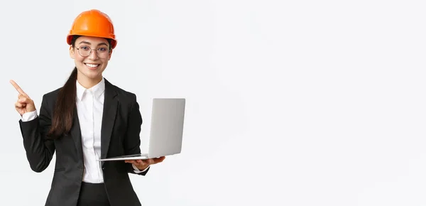 Usmívající se profesionální asijský ženský inženýr nebo architekt ve stavebnictví, nošení bezpečnostní přilby a skafandru, ukazující prstem vlevo při používání notebooku, stojící bílé pozadí — Stock fotografie