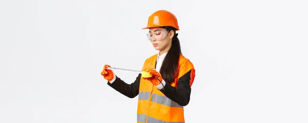 Serieuze Aziatische vrouwelijke bouwkundig ingenieur, technicus inspecteren lay-out, iets meten, kijken naar tape maatregel met gericht gezicht, staande over witte achtergrond in veiligheid uniform — Stockfoto