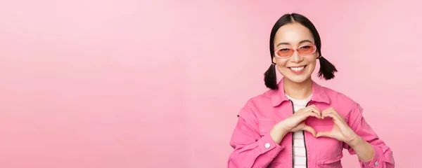 Bella coreano modello femminile in occhiali da sole alla moda, mostra il cuore, segno di cura, Ti amo gesto, si erge su sfondo rosa — Foto Stock