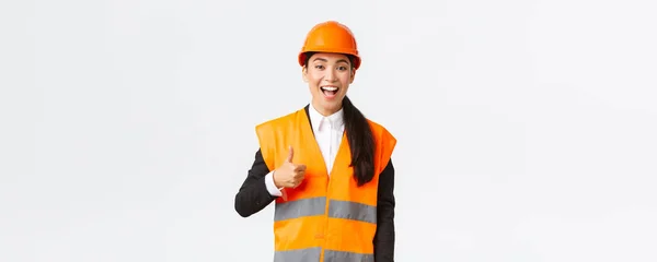 Soutien et heureux heureux asiatique femme ingénieur en chef dans le casque de sécurité montrant pouces levés dans l'approbation, satisfait des travaux de construction. Femme industrielle en veste réfléchissante inspecter la zone — Photo