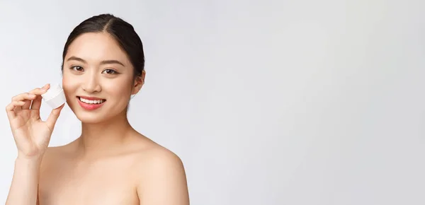 Hermosa joven mujer en blanco aislado fondo celebración cosmética cara crema, asiático —  Fotos de Stock