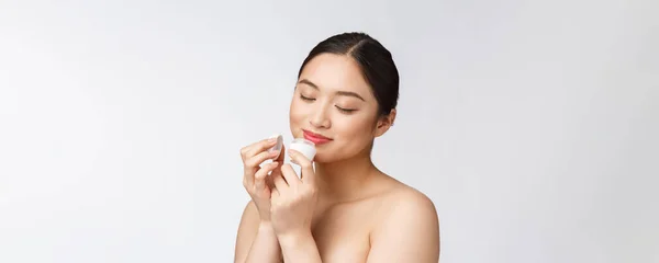 Krásná mladá žena na bílém izolovaném pozadí drží kosmetické obličeje krém, asijské — Stock fotografie