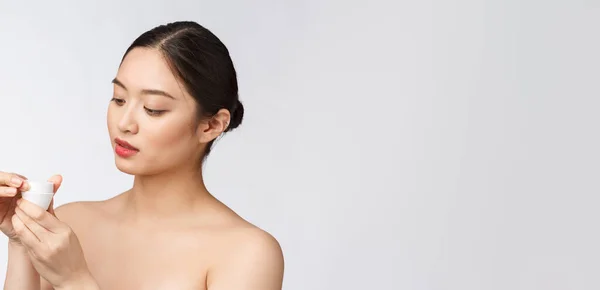 Krásná mladá žena na bílém izolovaném pozadí drží kosmetické obličeje krém, asijské — Stock fotografie