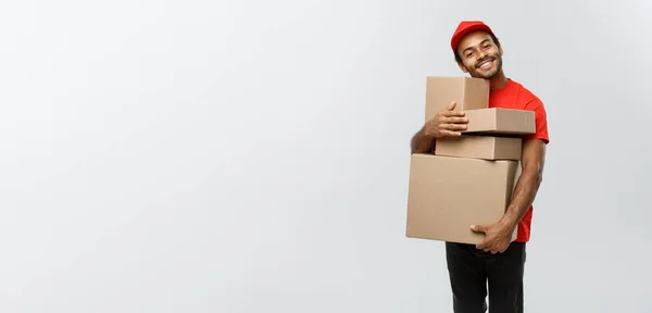 Lieferkonzept - Porträt eines glücklichen afroamerikanischen Zustellers in rotem Tuch, der ein Karton-Paket hält. Isoliert auf grauem Studiohintergrund. Kopierraum. — Stockfoto