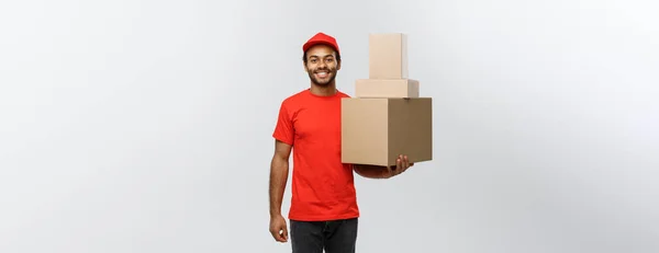 递送概念- -穿着红衣、提着盒子包裹的快乐非洲裔美国人送货员的肖像。在灰色工作室背景上被隔离。复制空间. — 图库照片