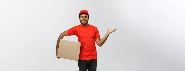 Lieferkonzept - Porträt eines glücklichen afrikanisch-amerikanischen Zustellers, der mit der Hand ein Karton-Paket präsentiert. isoliert auf grauem Studiohintergrund. Kopierraum. — Stockfoto