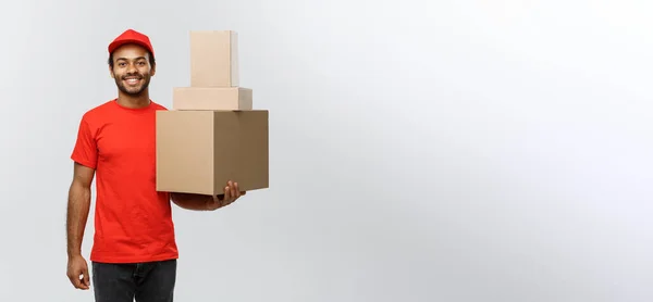 Concept de livraison - Portrait du livreur afro-américain heureux en tissu rouge tenant un paquet de boîte. Isolé sur fond de studio Grey. Espace de copie. — Photo