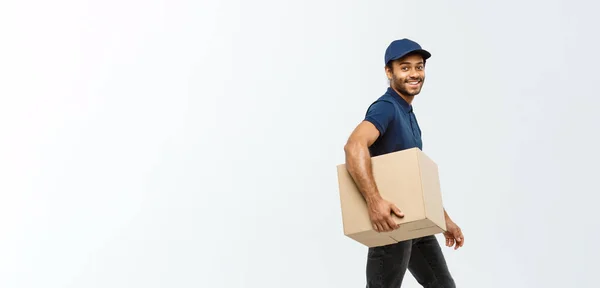 Conceito de entrega - Retrato de homem de entrega afro-americano feliz em pano azul andando para enviar um pacote de caixa para o cliente. Isolado no estúdio Grey Background. Espaço de cópia . — Fotografia de Stock