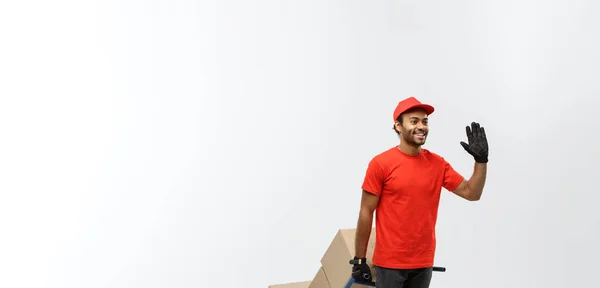 Concepto de entrega - Retrato del guapo repartidor afroamericano o mensajero empujando un camión de mano con una pila de cajas. Aislado en Grey studio Background. Copiar espacio . —  Fotos de Stock