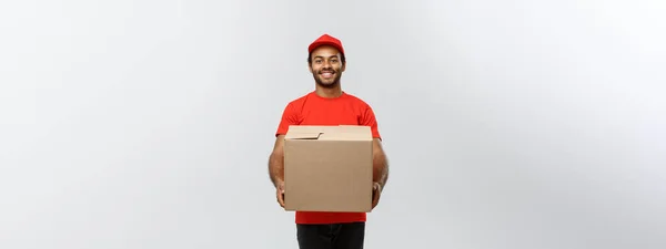 Conceito de entrega - Retrato de homem de entrega afro-americano feliz em pano vermelho segurando um pacote de caixa. Isolado no estúdio Grey Background. Espaço de cópia . — Fotografia de Stock