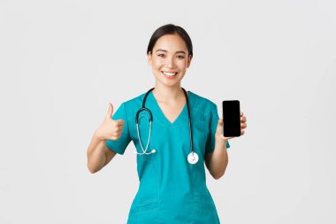 Covid-19, sağlık çalışanları ve online tıp konsepti. Genç Asyalı kadın doktor portresi, önlüklü doktor baş parmak ve akıllı telefon ekranı, tanıtım uygulaması, beyaz arka plan