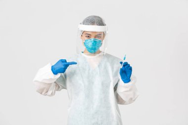 Covid-19, koronavirüs hastalığı, sağlık çalışanları konsepti. Ciddi bir Asyalı kadın doktor, PPE kişisel koruma ekipmanı doktoru, aşı şırıngasını işaret eden, beyaz arka plan