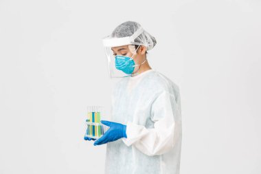 Covid-19, koronavirüs hastalığı, sağlık çalışanları konsepti. Dikkatli, ciddi Asyalı kadın doktor, teknoloji laboratuarı çalışanı kişisel koruma ekipmanları içinde elinde aşı tüpleri tutarken yan görüş.