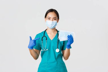 Covid-19, koronavirüs hastalığı, sağlık çalışanları konsepti. Gülümseyen Asyalı doktor, doktor veya hemşire kauçuk eldivenli ve önlüklü hasta güvenliği için tıbbi maskeleri işaret ediyor.