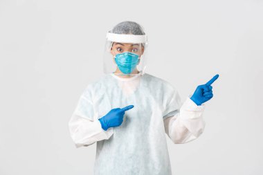 Covid-19, koronavirüs hastalığı, sağlık çalışanları konsepti. Şaşırmış ve şaşırmış Asyalı kadın doktor, kişisel koruma ekipmanları içinde teknoloji laboratuarı çalışanı şaşırmış ve parmağıyla işaret ediyor.