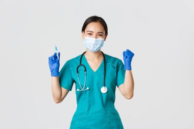 Covid-19, koronavirüs hastalığı, sağlık çalışanları konsepti. Neşeli zafer kazanan Asyalı kadın doktor, tıbbi maske ve eldiven takan doktor, aşıyla şırınga tutarken seviniyor.