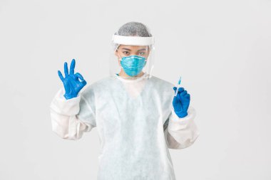 Covid-19, koronavirüs hastalığı, sağlık çalışanları konsepti. Kişisel koruma ekipmanlarında kendine güvenen ciddi Asyalı kadın doktor, iyi bir jest göster, aşıyla şırıngayı tut, beyaz arka plan
