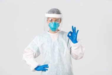 Covid-19, koronavirüs hastalığı, sağlık çalışanları konsepti. Kişisel koruma ekipmanları içinde ciddi görünümlü Asyalı kadın doktor, iyi bir jest gösteriyor, hastanın güvenliğini sağlıyor.
