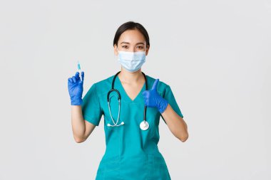 Covid-19, koronavirüs hastalığı, sağlık çalışanları konsepti. Hoş gülümseyen Asyalı kadın doktor, tıbbi maske ve eldiven giyen doktor, baş parmağını kaldır, aşı ile şırınga tut.