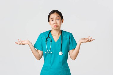 Covid-19, sağlık çalışanları, salgın konsepti. Cahil Asyalı kadın hemşire portresi, omuz silkip yan yana uzanan kadın doktor, ne bileyim, yardım edemem, beyaz arka plan.