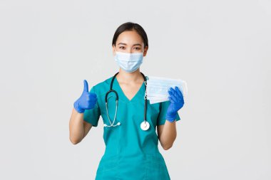 Covid-19, koronavirüs hastalığı, sağlık çalışanları konsepti. Gülümseyen Asyalı doktor, önlüklü hemşire ve lastik eldiven, baş parmak ve tıbbi maskeler, beyaz arka plan