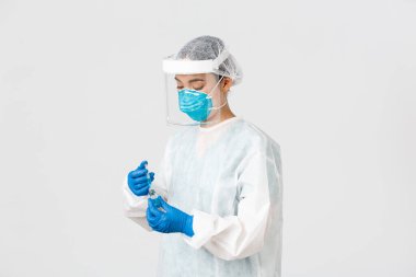 Covid-19, koronavirüs hastalığı, sağlık çalışanları konsepti. Ciddi görünümlü kadın hekim, kişisel koruma ekipmanlarında Asyalı doktor aşıyla birlikte ampul içine şırınga yerleştirin.