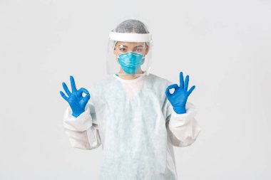 Covid-19, koronavirüs hastalığı, sağlık çalışanları konsepti. Kendinden emin ve ciddi Asyalı kadın doktor, kişisel koruma ekipmanlarında laboratuvar teknisyeni onaylı jest gösteriyor.