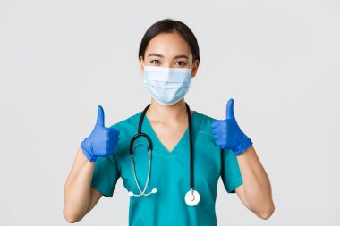 Covid-19, koronavirüs hastalığı, sağlık çalışanları konsepti. Kendinden emin gülümseyen Asyalı doktor, tıbbi maske takan hemşire ve lastik eldiven, onaylama için onay ver, hizmet et, bilgi topla.