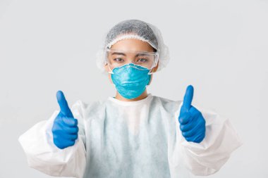 Covid-19, koronavirüs hastalığı, sağlık çalışanları konsepti. Şahsi koruma ekipmanlarıyla kendine güvenen Asyalı kadın doktorların yakın plan fotoğrafları. Başparmak kaldırma, tavsiye etme veya kalite güvencesi