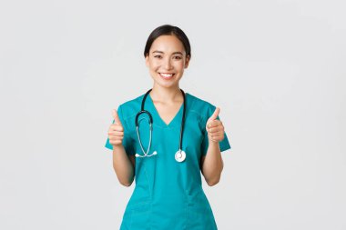 Covid-19, sağlık çalışanları, salgın konsepti. Profesyonel kendine güvenen doktor, önlüklü kadın doktor emin ifadeyle baş parmağını kaldırıyor, her şeyin iyi, mükemmel ya da mükemmel olduğundan emin oluyor.