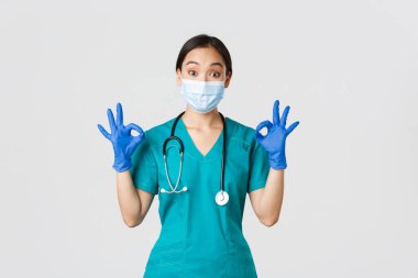 Covid-19, koronavirüs hastalığı, sağlık çalışanları konsepti. Etkilenmiş Asyalı kadın doktor, tıbbi maskeli doktor, kauçuk küreler ve önlükler. Tamam, jest, iltifat, övgü, mükemmel iş.