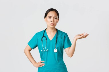 Covid-19, healthcare workers, pandemic concept. Frustrated and annoyed asian female doctor, nurse in scrubs raise one hand and looking questioned and pissed-off, complaining something