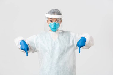 Covid-19, koronavirüs hastalığı, sağlık çalışanları konsepti. Şaşırmış ve heyecanlı kadın doktor, kişisel koruyucu ekipman hekimi parmakları aşağıda, beyaz arka planda duruyor.