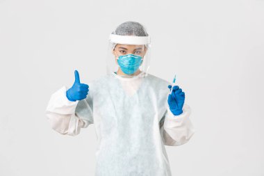 Covid-19, koronavirüs hastalığı, sağlık çalışanları konsepti. Kişisel koruma ekipmanlarında kendine güvenen ciddi Asyalı kadın doktor, baş parmağını kaldır ve aşı ile şırıngayı tut, beyaz arka plan